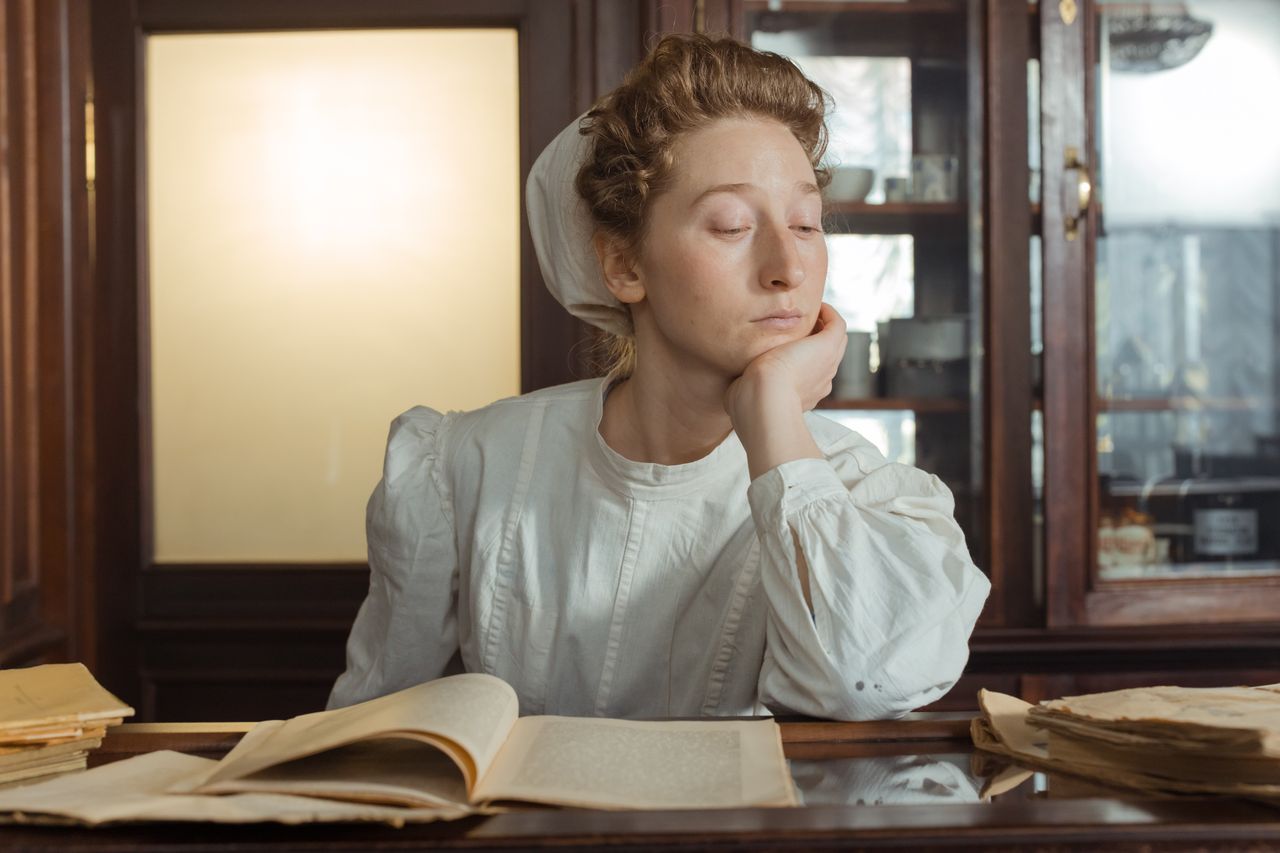 Woman in vintage attire sitting indoors, reading an open book with an introspective expression.